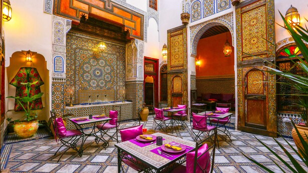 A Moroccan-style interior with ornate tile work, vibrant seating, and a central fountain. - Dar Attajalli (Fes)