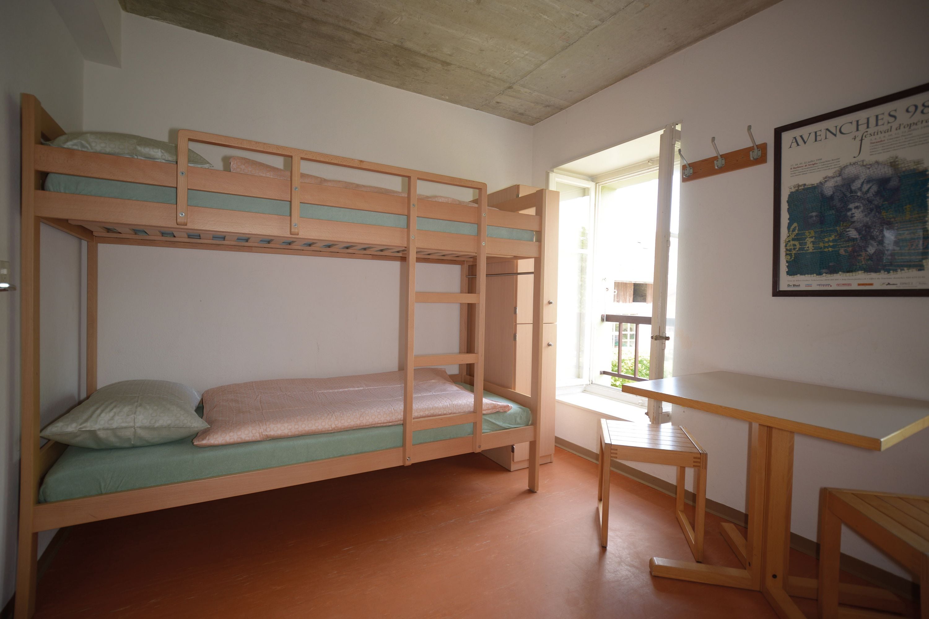 Ein Stockbett mit Schreibtisch und Stuhl, ein Poster an der Wand und ein Fenster mit Blick ins Freie.