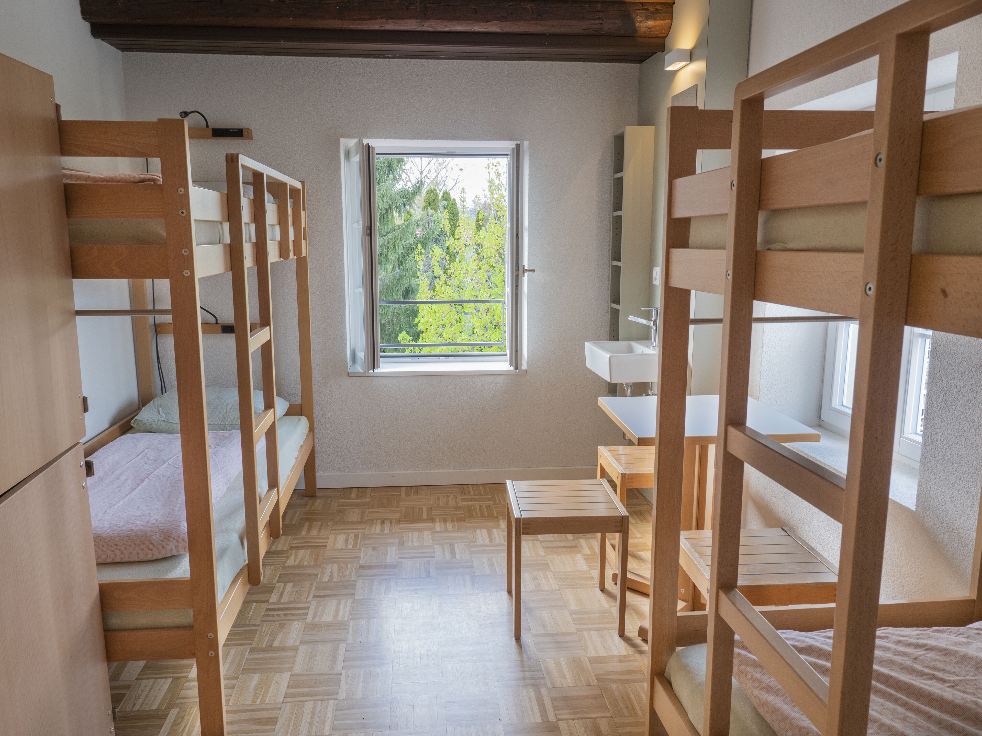 A dormitory room with bunk beds, a small table, a sink, and a window with a view of greenery.