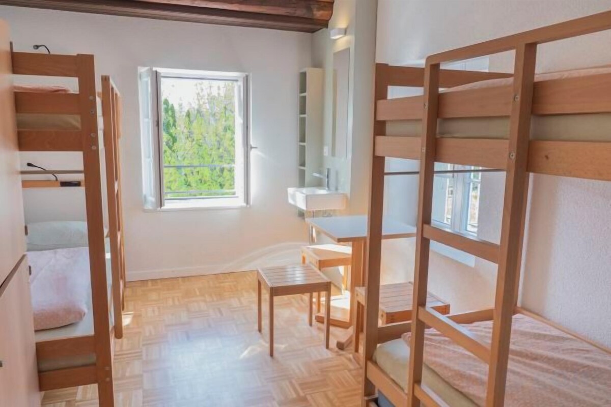Ein Zimmer mit Etagenbetten, einem kleinen Tisch und einem Fenster mit Blick auf Grünflächen.
