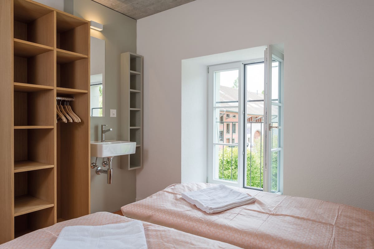 A bedroom with a large window, a wooden wardrobe, and a bathroom with a sink.