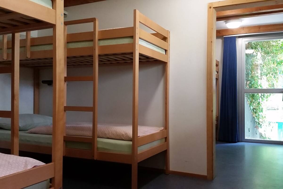 A room with bunk beds, a window with a view of greenery, and a door leading to another room.