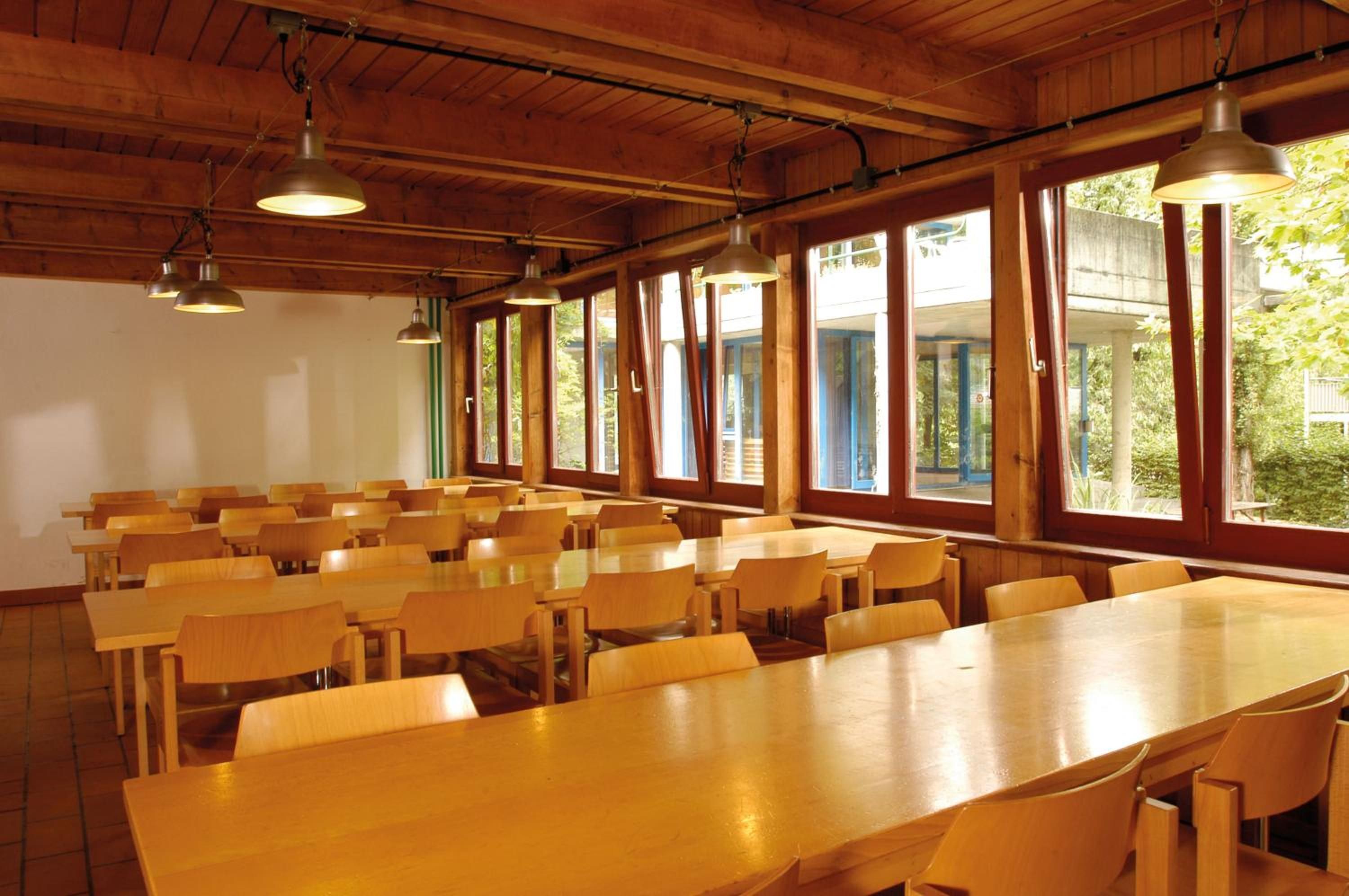 A large hall with wooden tables and chairs, high ceilings, and large windows.