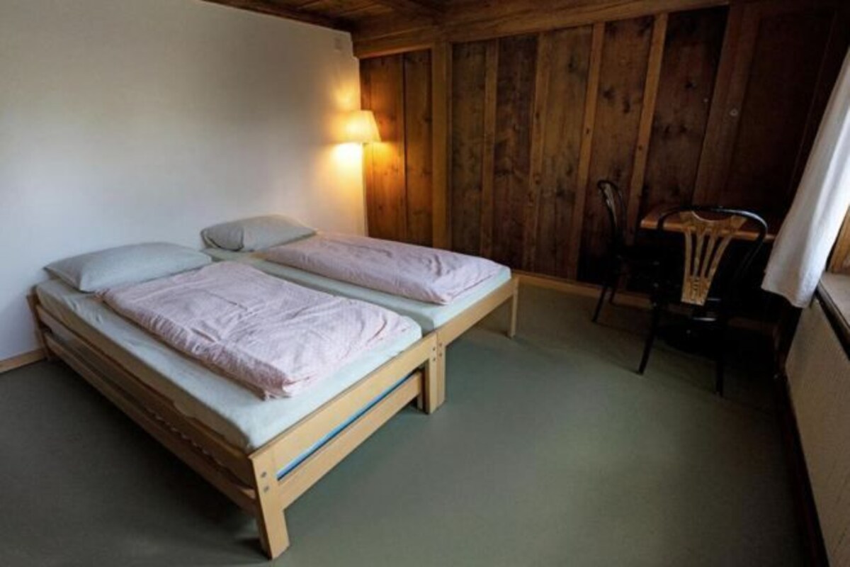 Una habitación pequeña y acogedora con dos camas individuales, una silla de madera y una lámpara de pared.