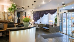 Een moderne hotellobby met een receptiebalie, een zithoek en grote ramen.
