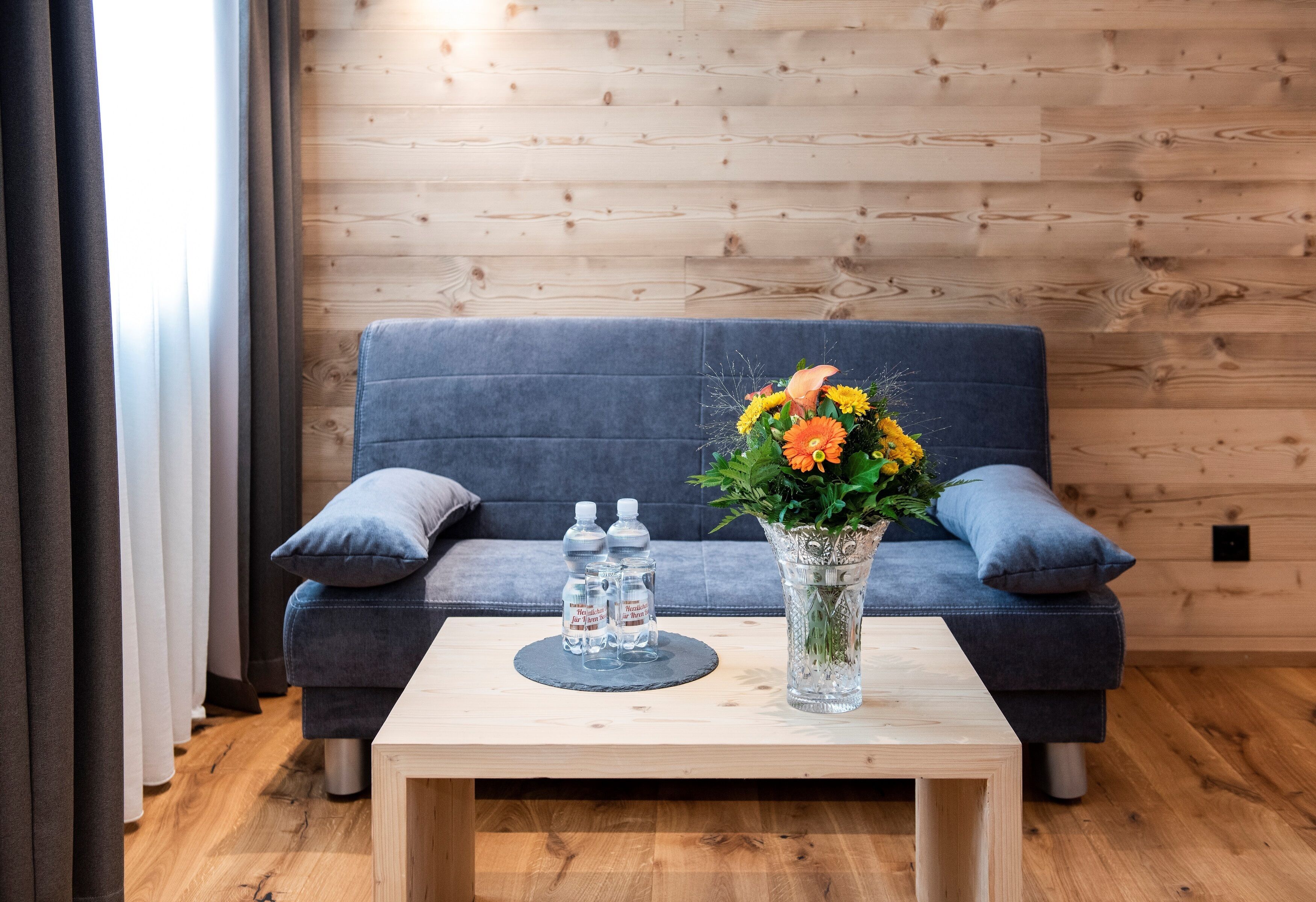 A modern room with a wooden floor, a dark grey sofa, a small wooden coffee table, a vase with flowers, and two water bottles.