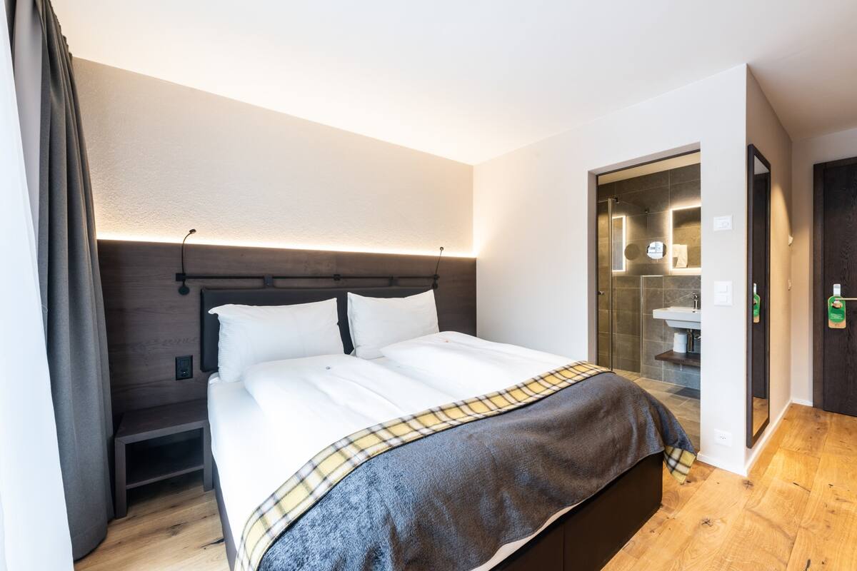 Une chambre d’hôtel moderne dotée d’un grand lit, d’une table de chevet, d’une petite commode, d’une salle de bain avec une douche en verre et d’un plancher en bois.
