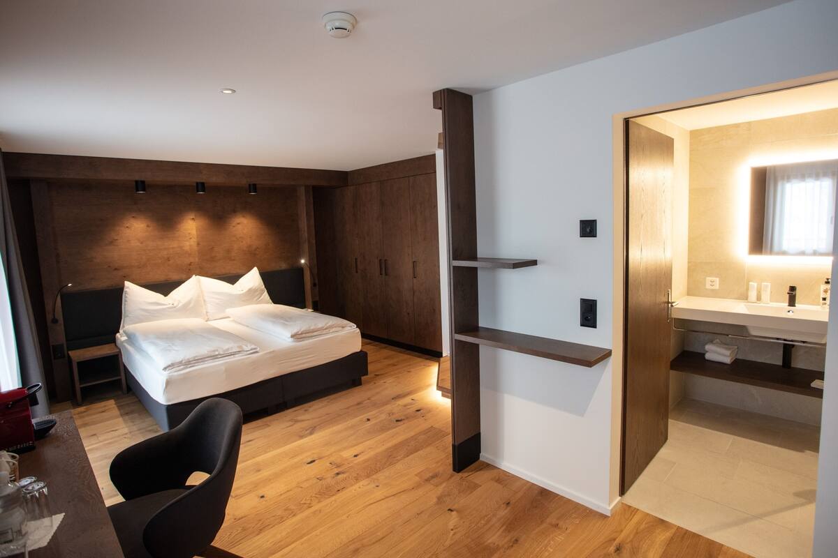 Une chambre d’hôtel moderne avec un plancher en bois, un lit avec du linge de maison blanc, un bureau avec une chaise, une armoire intégrée et une salle de bain avec un miroir et un lavabo.