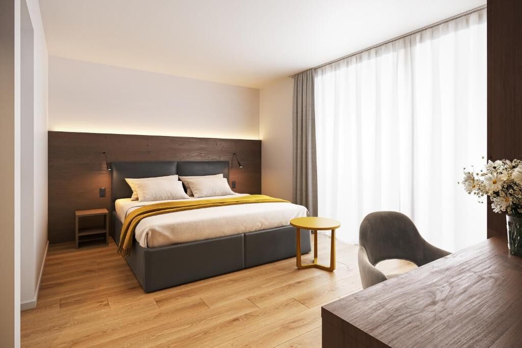 A modern hotel room with a large bed, a small side table, a desk with a chair, and a large window with curtains.
