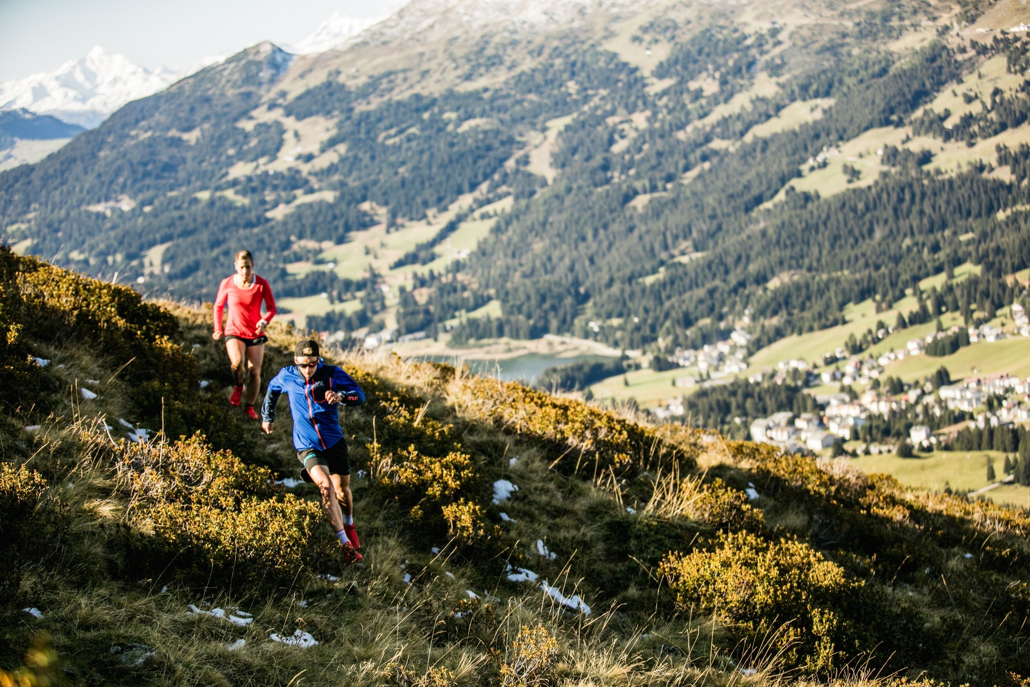 Two people running on a grassy hill with a mountainous landscape in the background.