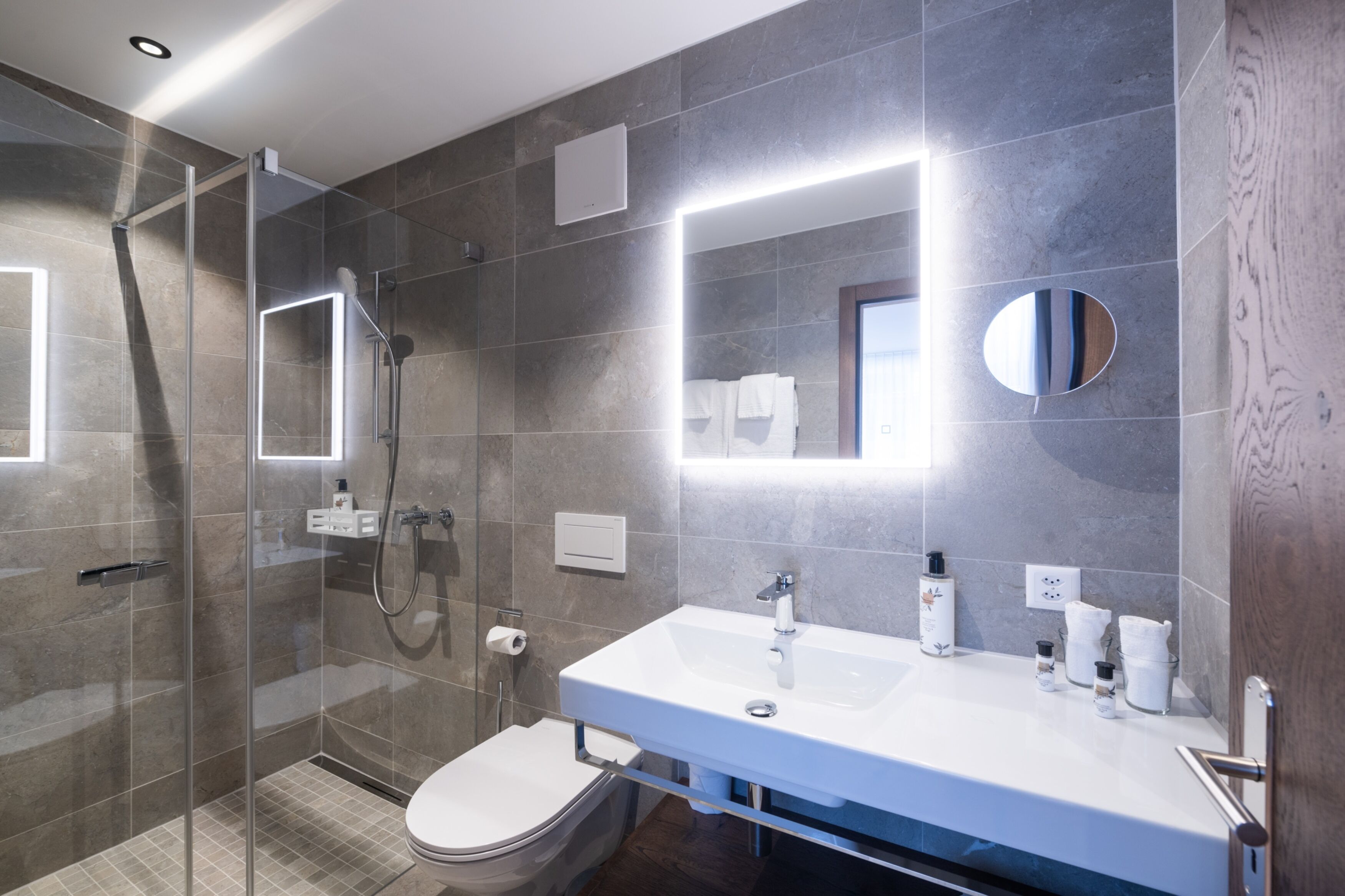 A modern bathroom with a glass-enclosed shower, a white sink with toiletries, and a large mirror.