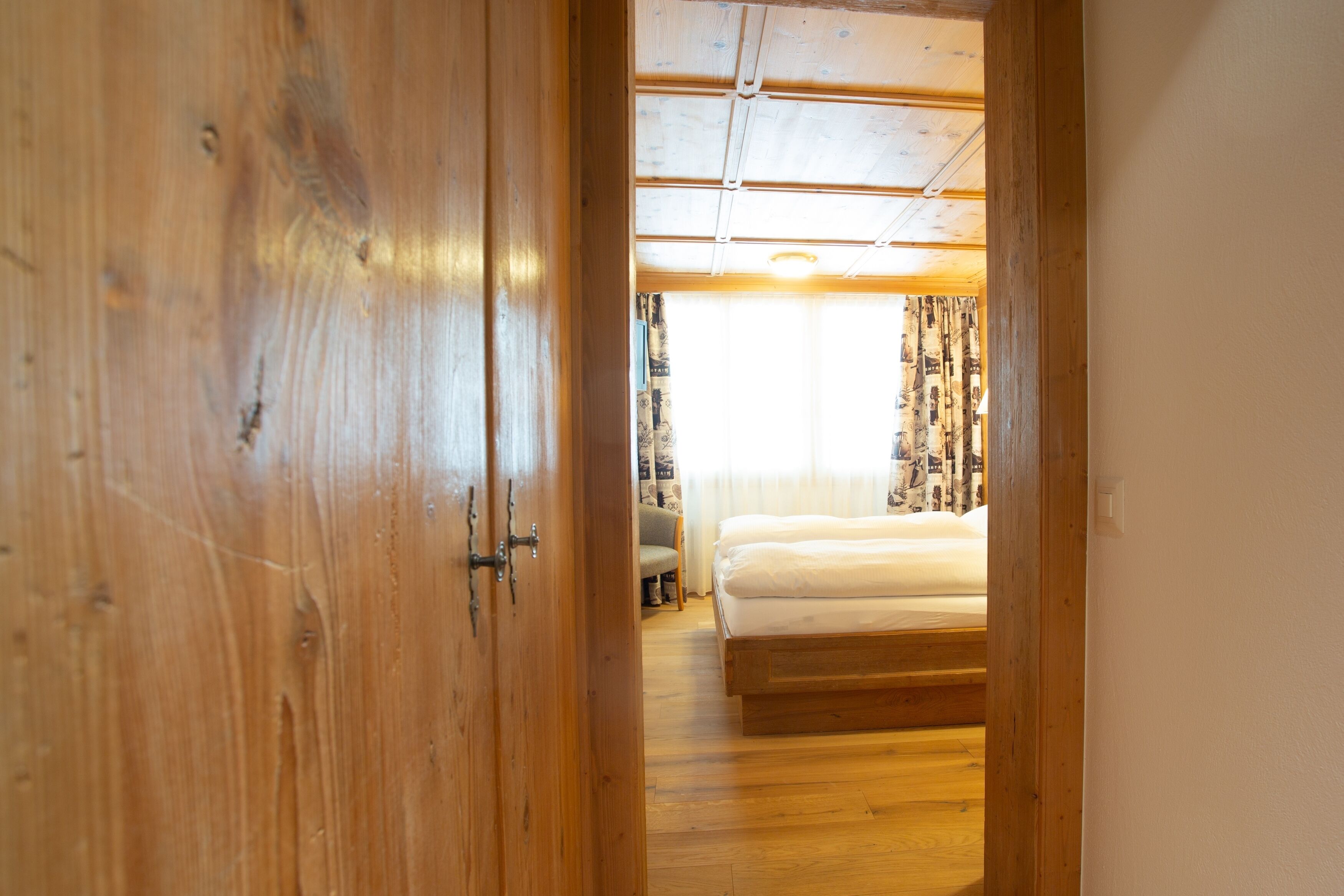 A wooden door leading to a bedroom with a bed, a ceiling fan, and a window with curtains.