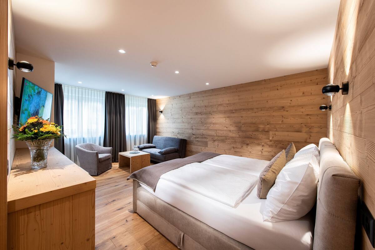 Une chambre d’hôtel moderne avec un mur en bois, un grand lit avec du linge de maison blanc, un canapé gris, une petite table et un téléviseur posé sur une console en bois.