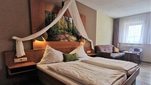 A hotel room with a large bed, a nightstand, a chair, a small table, and a painting on the wall. - Hotel Drei Lilien (Werbach)
