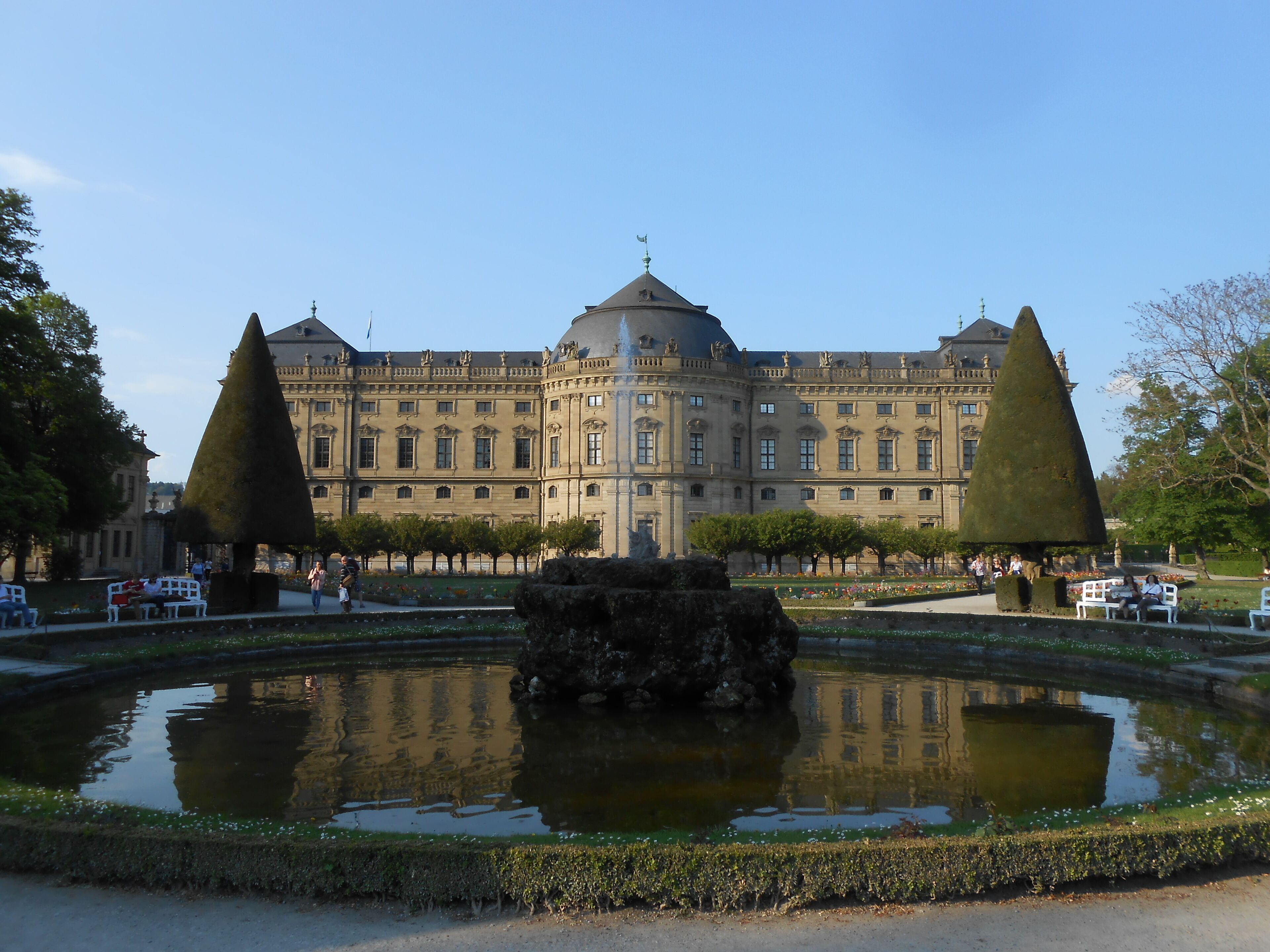Un edificio grande e histórico con arquitectura simétrica, una fuente central y un jardín bien cuidado con árboles podados.