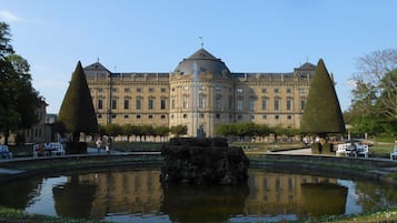 Un edificio grande e histórico con arquitectura simétrica, una fuente central y un jardín bien cuidado con árboles podados.