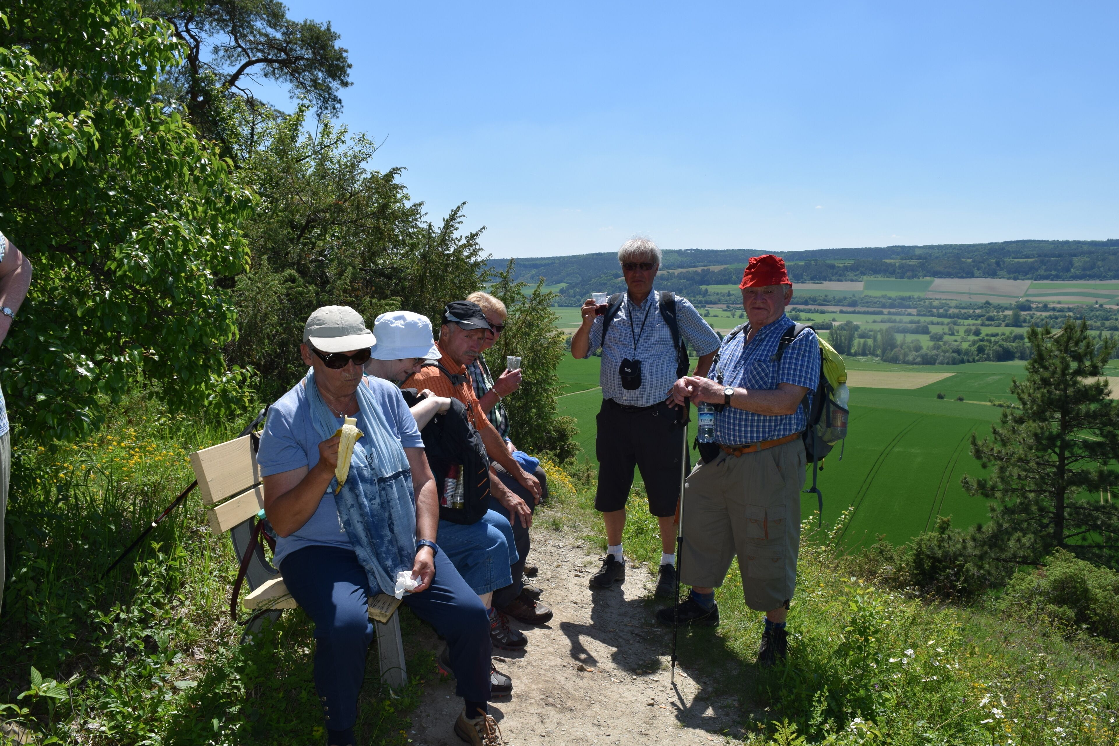 Un gruppo di persone sedute su una panchina e in piedi su un sentiero, con vista panoramica su campi verdi e alberi.