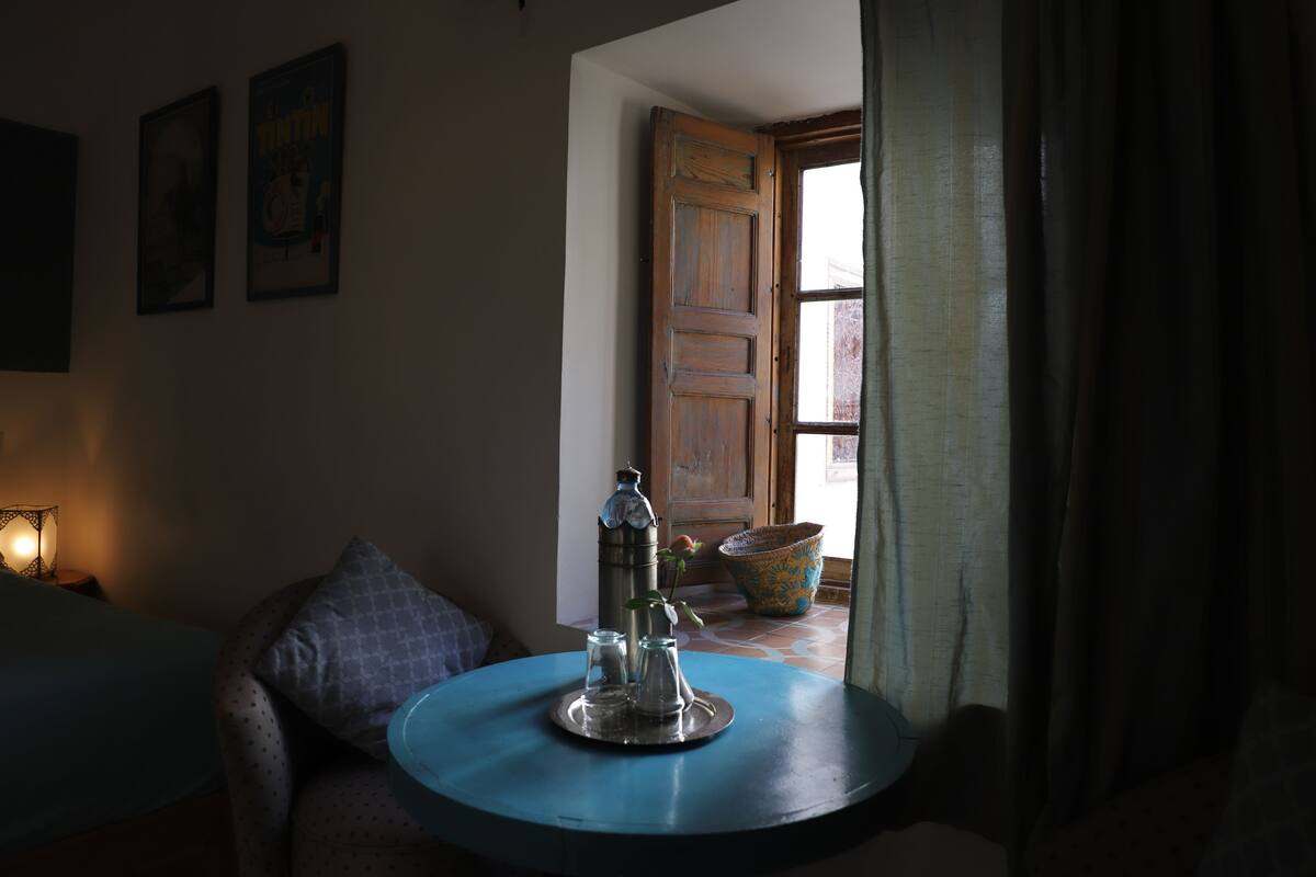 A room with a round blue table, a chair, a lamp, and a door leading outside.
