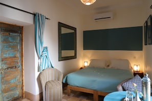A bedroom with a wooden bed, a striped chair, a blue bedspread, and a wall-mounted air conditioner. - Riad le Coq Fou (Marrakech)