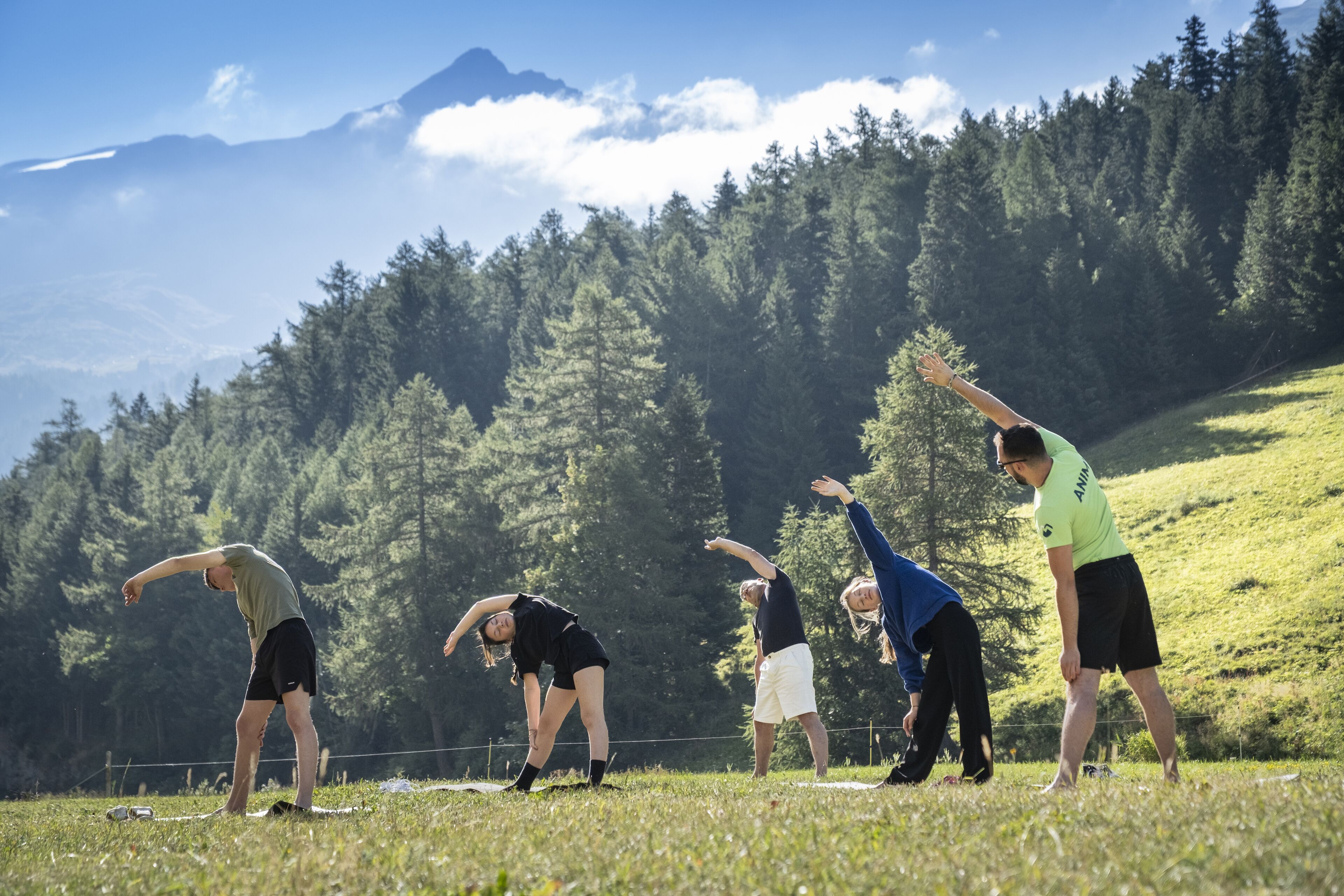 A group of people performing a stretching exercise outdoors with a mountainous backdrop.