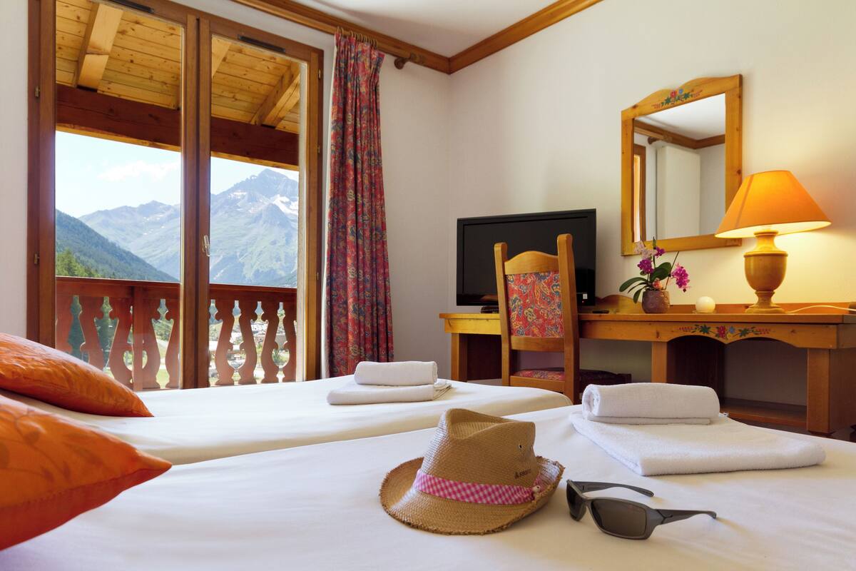 A hotel room with two beds, a desk with a computer, a chair, a lamp, and a view of mountains.