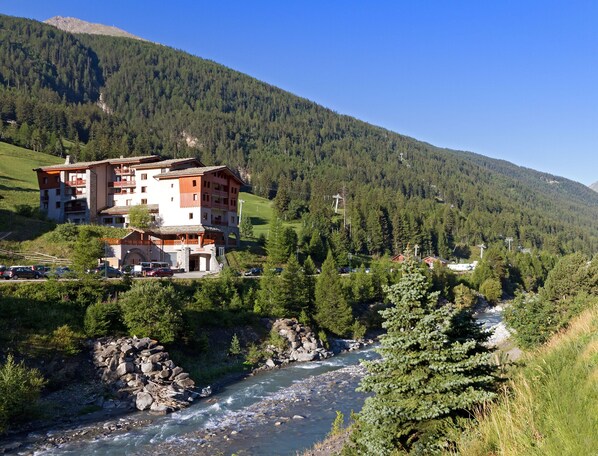 Un hôtel à plusieurs étages doté de balcons, avec une rivière au premier plan et une chaîne de montagnes en arrière-plan.