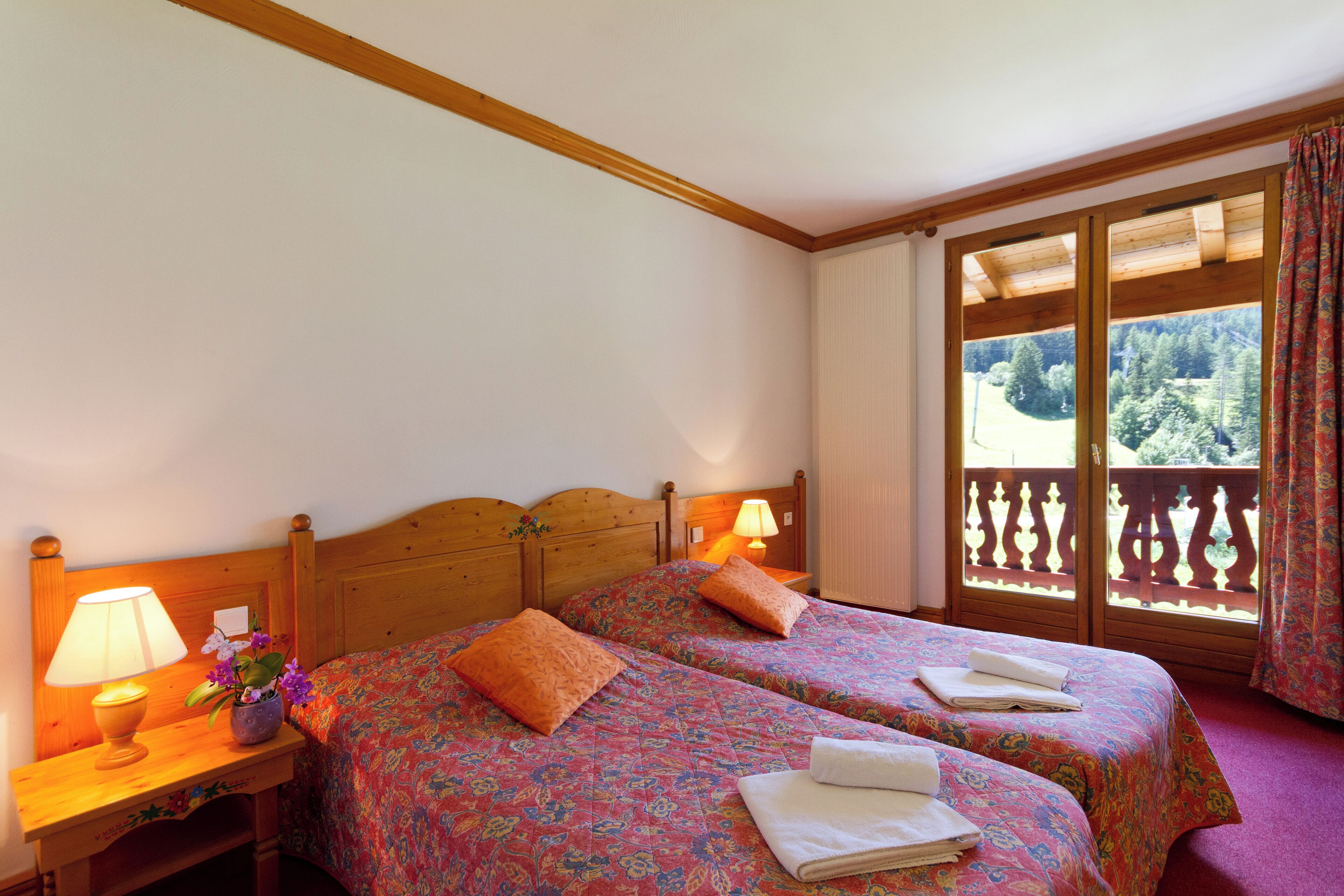 Camera d'albergo con due letti, testiere in legno, un comodino con una lampada e una finestra vista alberi.