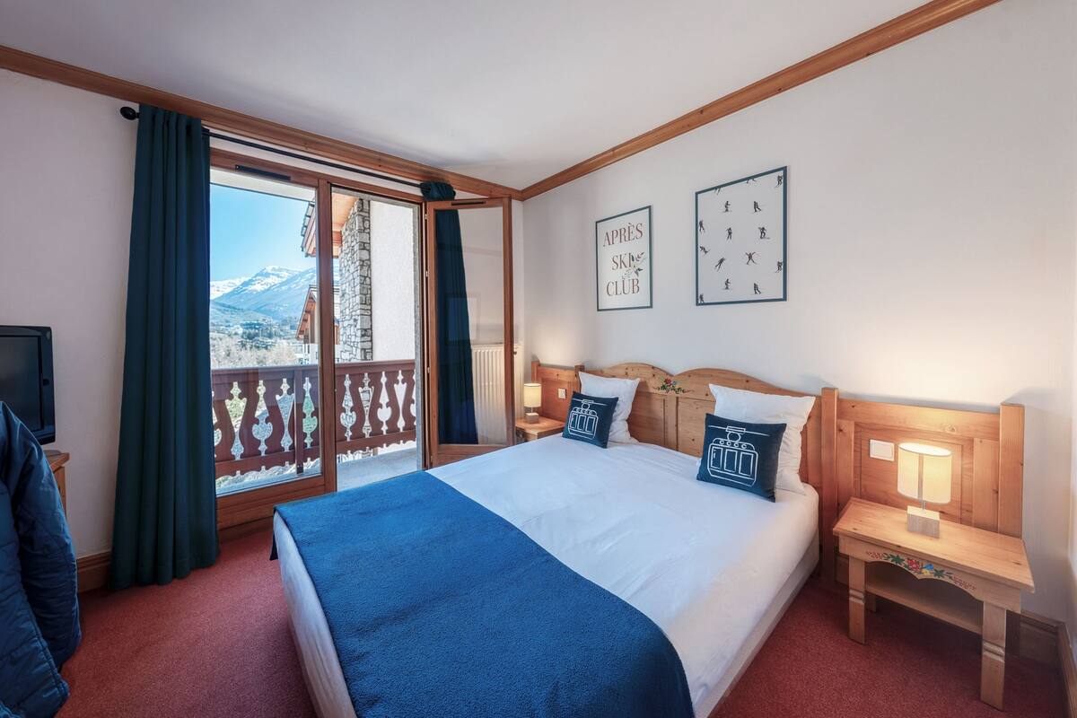 A hotel room with a bed, a nightstand, a lamp, a TV, a window with curtains, and a view of mountains.