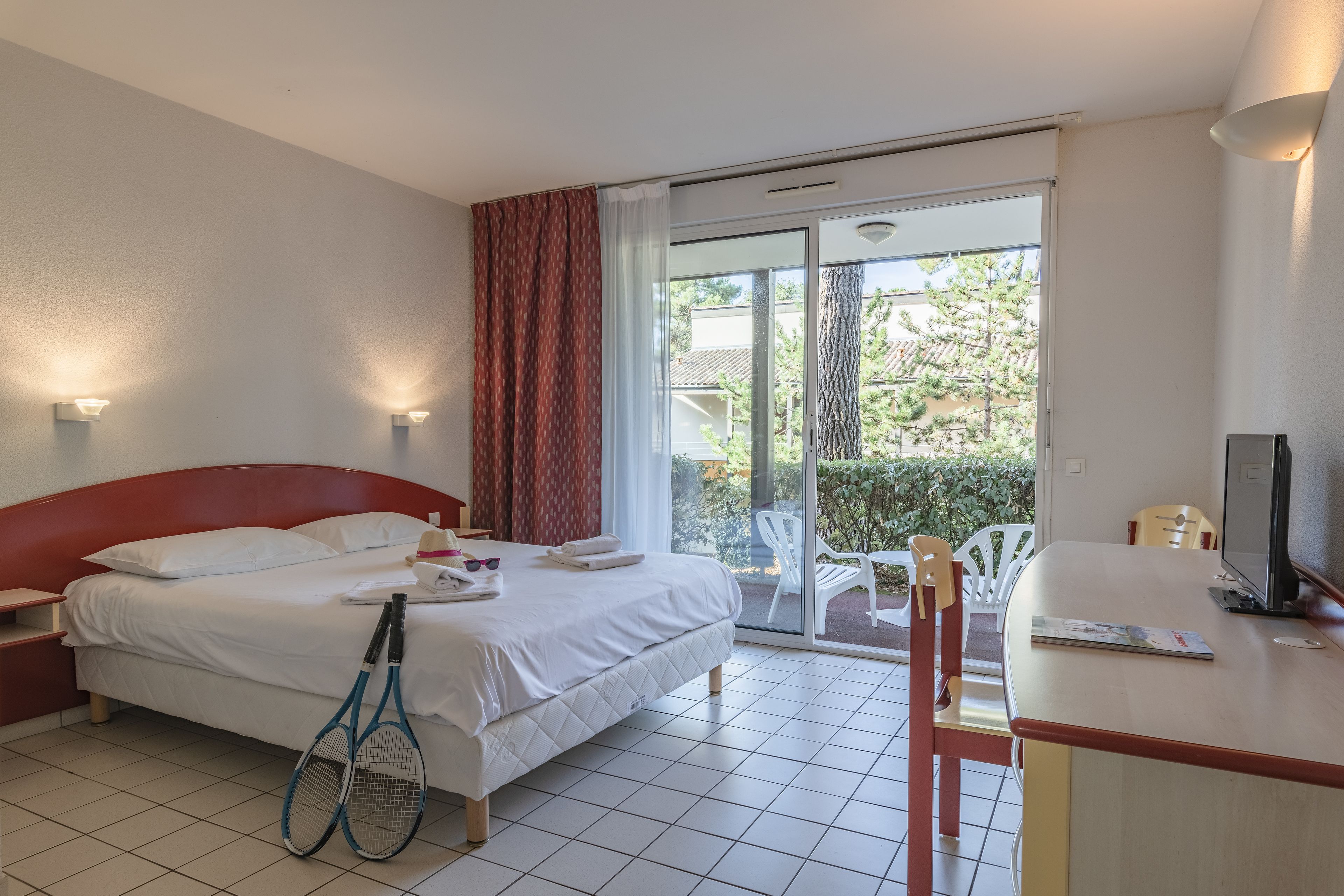 Ein Hotelzimmer mit Bett, Schreibtisch mit Stuhl, Fernseher und Balkon mit Blick auf Bäume und Gebäude.