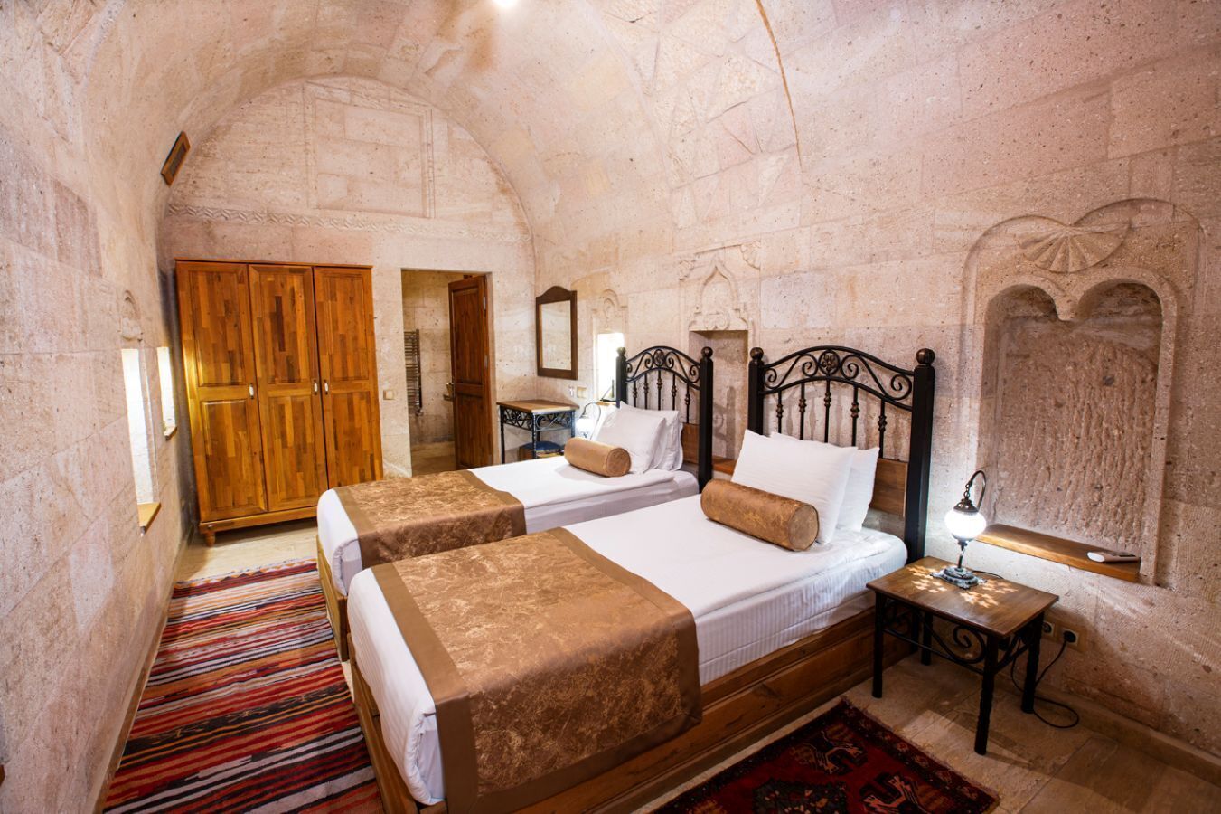 A room with two beds, wooden wardrobe, and a small table with a lamp.