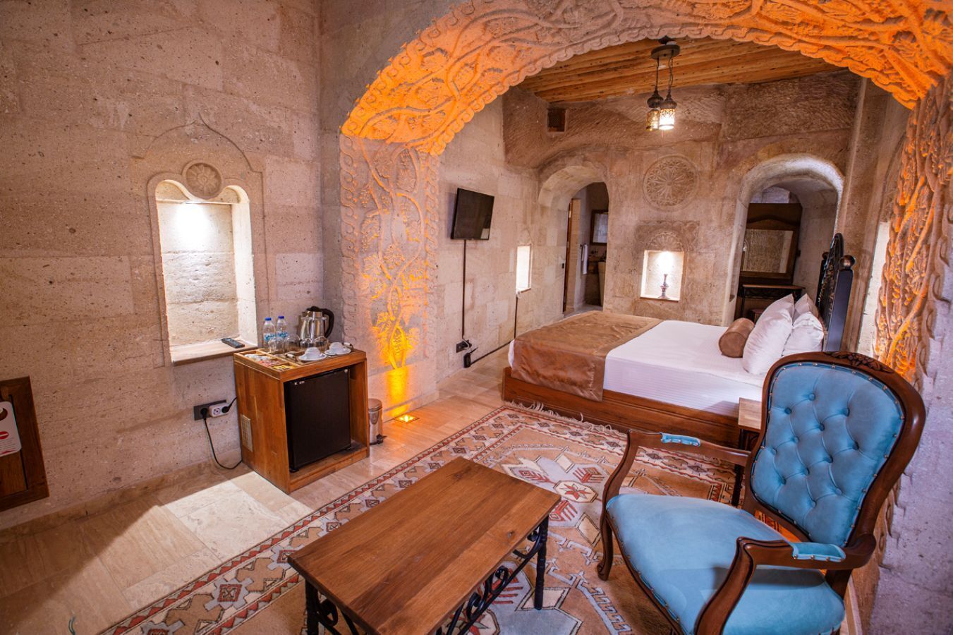 A room with a bed, a wooden desk, a chair, and a small table. There is a wall-mounted TV and a decorative light fixture.