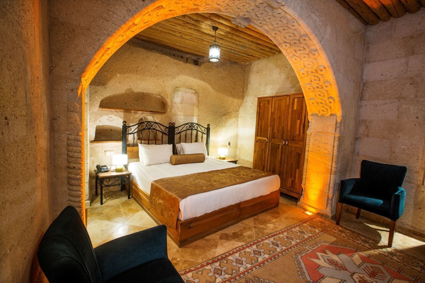 A cozy bedroom with a large bed, a wooden wardrobe, a chair, and a patterned rug.