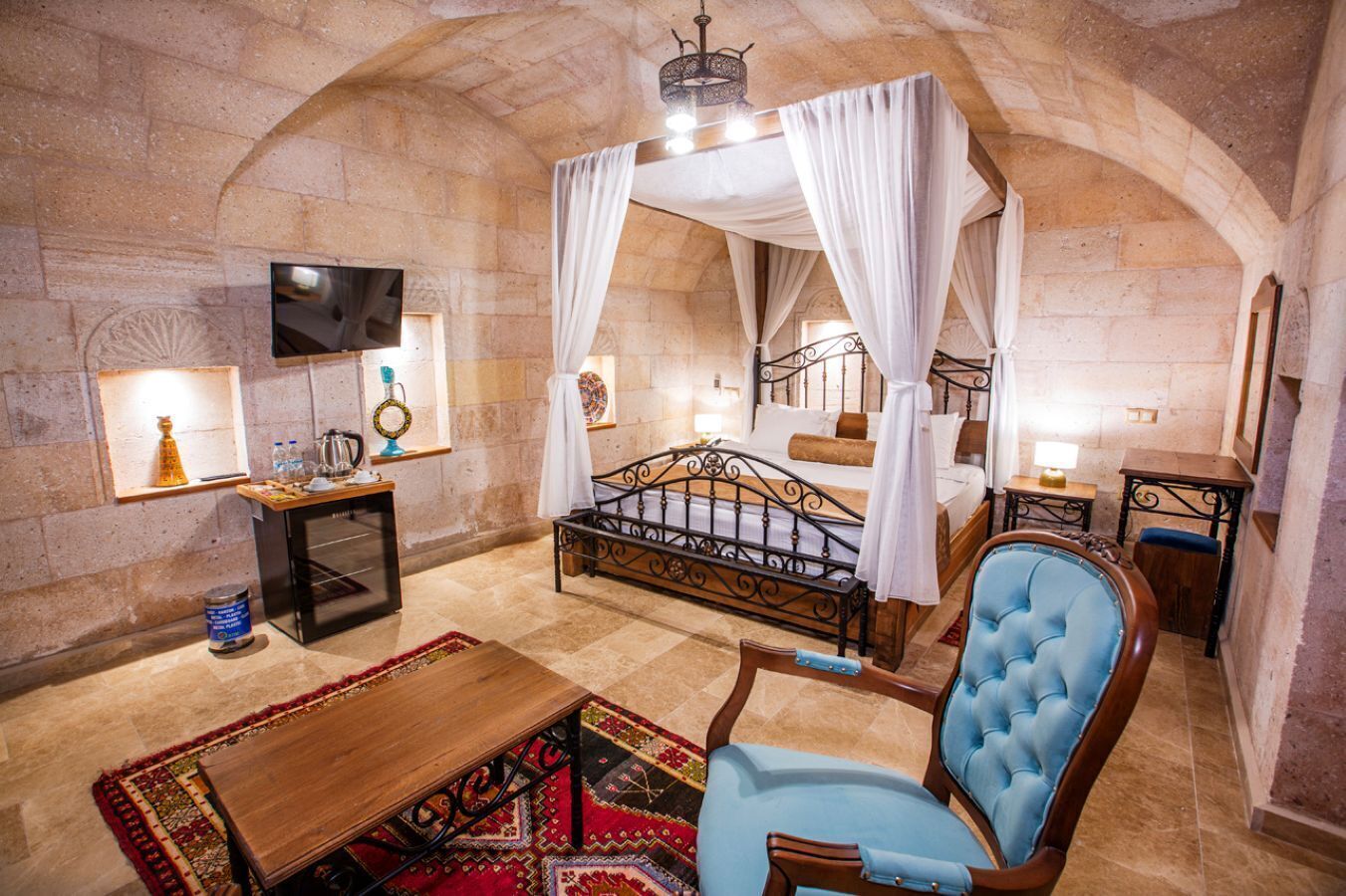 A room with a four-poster bed, a wooden table, a blue armchair, and a small desk with a lamp.