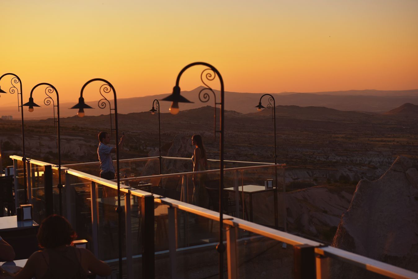 A terrace with metal railings and hanging lamps overlooking a desert landscape during sunset.