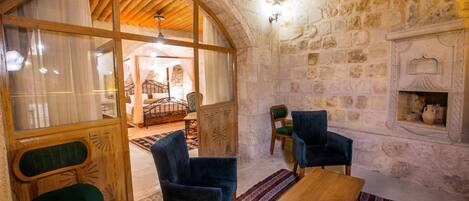 A rustic room with a stone wall, a wooden ceiling, a bed with a canopy, two blue chairs, a wooden table, and a patterned rug.