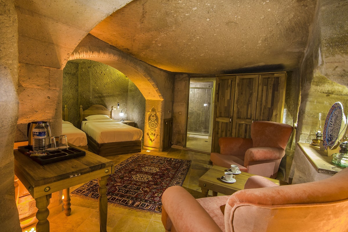 Een kamer met een stenen boog, een bed, een zithoek met een stoel, een bijzettafeltje en uitzicht door een raam.