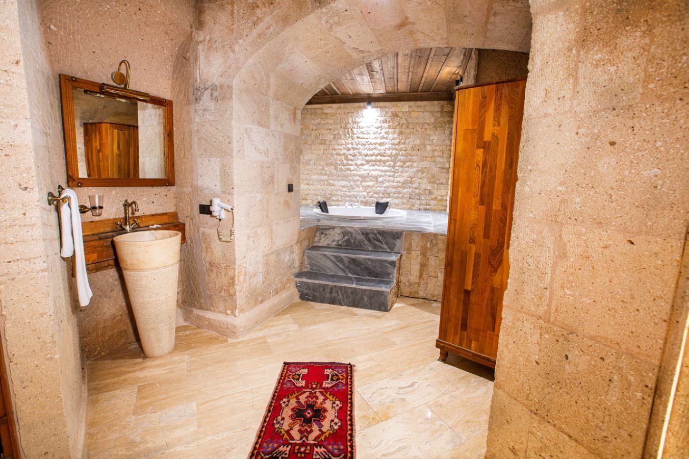 A bathroom with a stone sink, a wooden vanity, a marble-topped bathtub, and a red patterned rug.