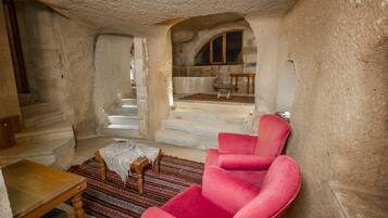 Una habitación con un sillón rojo, una pequeña mesa de madera y un tapete estampado. Se ve una escalera que conduce a un nivel superior con una ventana y una mesa pequeña.