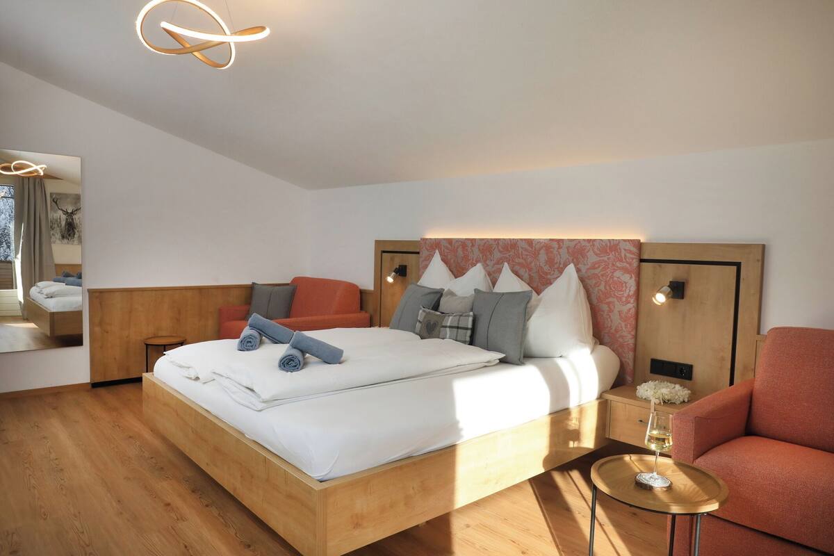 Et moderne hotelværelse med en træseng, en rød lænestol, et lille rundt bord med et glas vin og et spejl der reflekterer rummet.