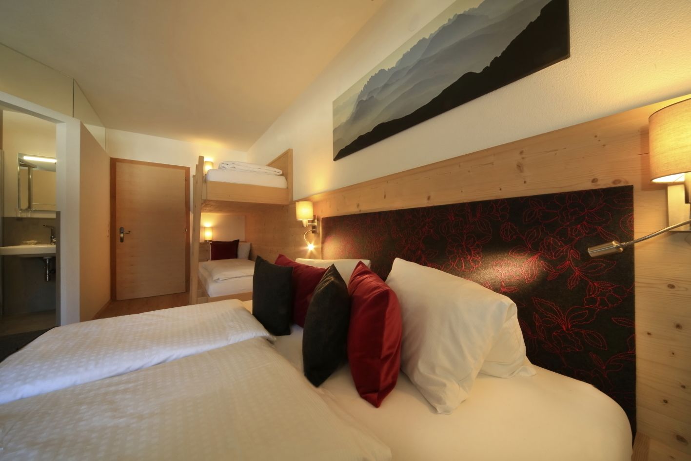 A hotel room with a large bed, a wooden headboard, and a painting above the bed. There is a bathroom visible in the background.