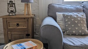 A cozy room with a grey sofa, a wooden nightstand with a lamp, and a tray with a book titled "The Heart Keeper". - Humphries Cross Apartments (Rye)