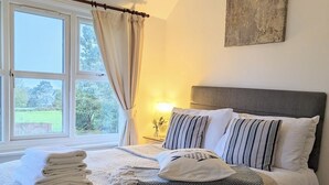 A neatly made bed with a striped pillow, a bedside lamp, and a view of the outdoors through a window with curtains. - Humphries Cross Apartments (Rye)