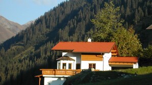 Un chalet à deux étages, avec un toit recouvert de tuiles rouges, un balcon en bois et une vue sur une chaîne de montagnes boisées.