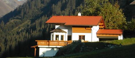 A two-story chalet with a red-tiled roof, wooden balcony, and a view of a forested mountain range.