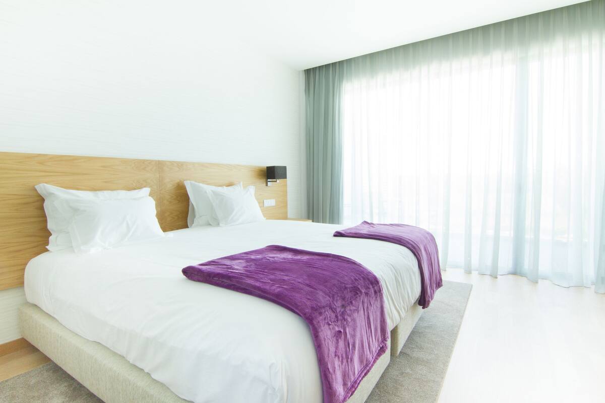 A modern hotel room with a large bed, a wooden headboard, and a large window with sheer curtains.