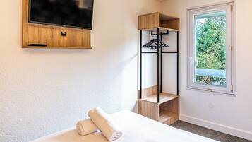 Habitación de hotel con cama, televisor montado en un panel de madera, estante de madera con perchero y ventana con vista a follaje.