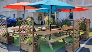Un área de patio con mesas de picnic y sombrillas, rodeada de macetas de madera con flores.
