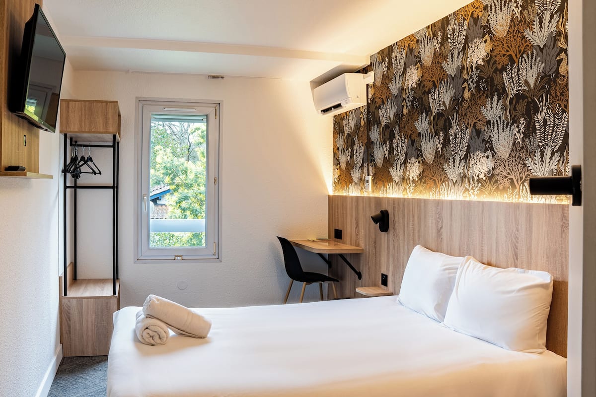 Habitació d'hotel amb un llit, escriptori, cadira, televisió i finestra amb vista a arbres.