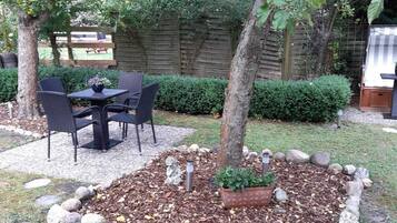 Een terras met een tafel en stoelen, omgeven door een bloembed met mulch en stenen, en een houten hek op de achtergrond.