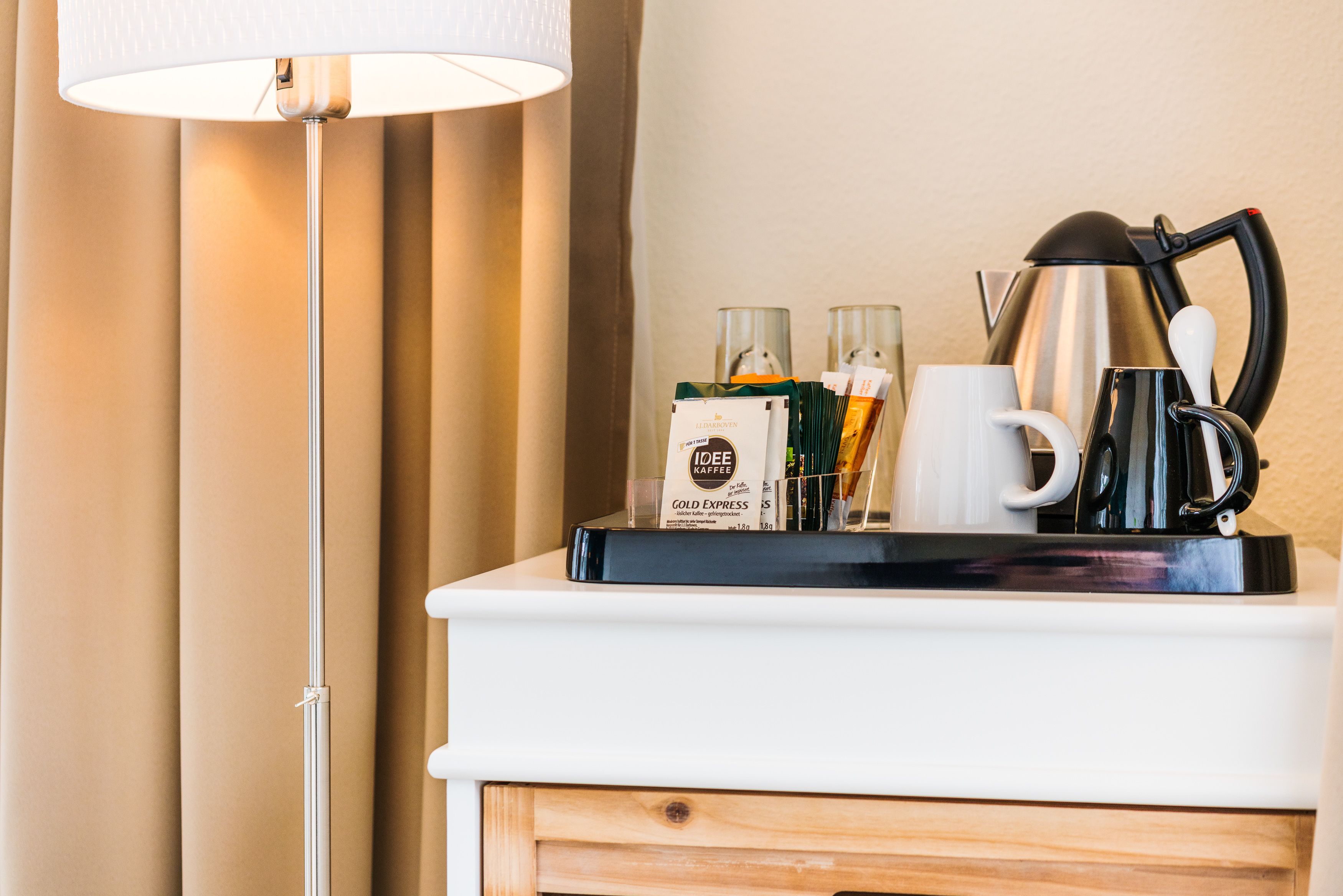 A hotel room with a white bedside table, a lamp, a coffee maker, and a tray with coffee packets and a mug.